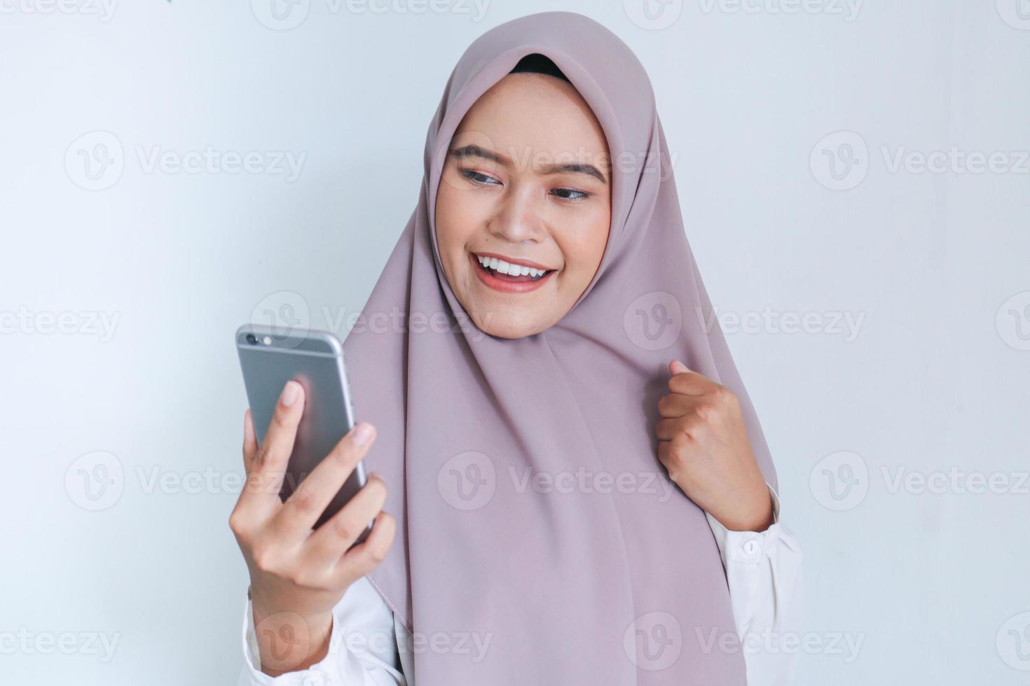 la giovane donna asiatica dell'islam che indossa il velo è felice ed entusiasta di celebrare in ciò che vede sullo smartphone. donna indonesiana su sfondo grigio foto