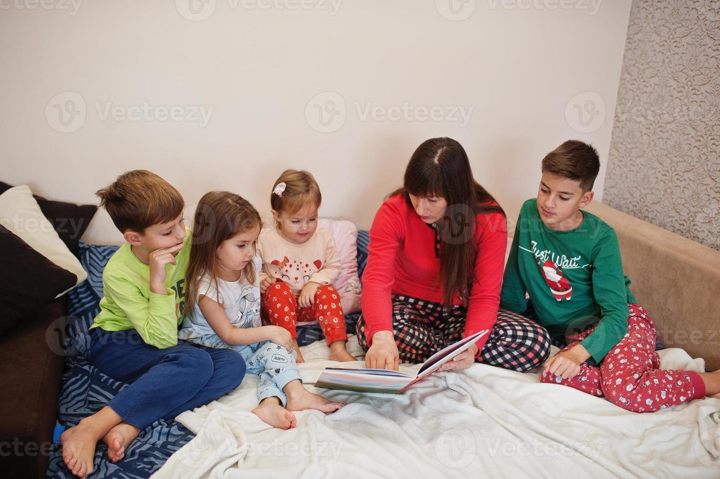 la grande famiglia felice si diverte insieme in camera da letto. concetto di mattina in famiglia numerosa. la madre con quattro bambini indossa il pigiama legge il libro a letto a casa. foto