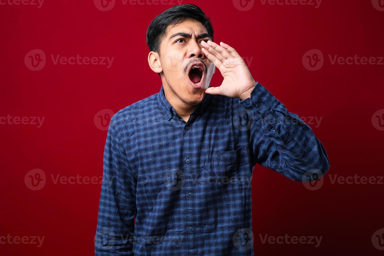 giovane uomo asiatico che indossa una camicia casual in stile gridando e urlando ad alta voce di lato con la mano sulla bocca foto