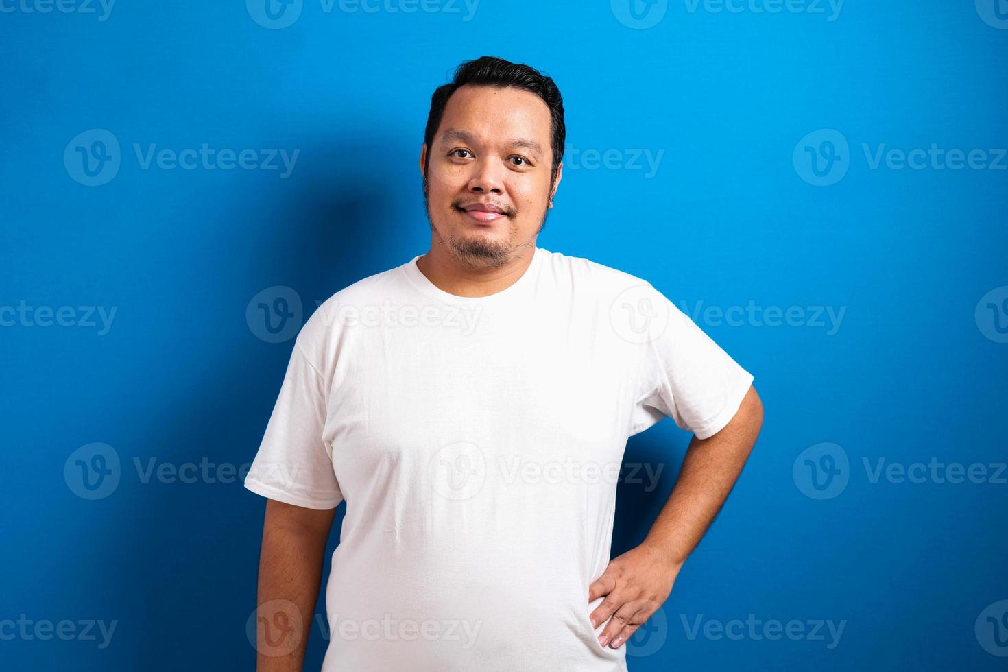 ragazzo asiatico grasso che indossa una t-shirt bianca si alza mettendo le mani sulla sua vita sorridendo verso la telecamera. l'uomo mostra un gesto sicuro di sé, su sfondo blu. foto