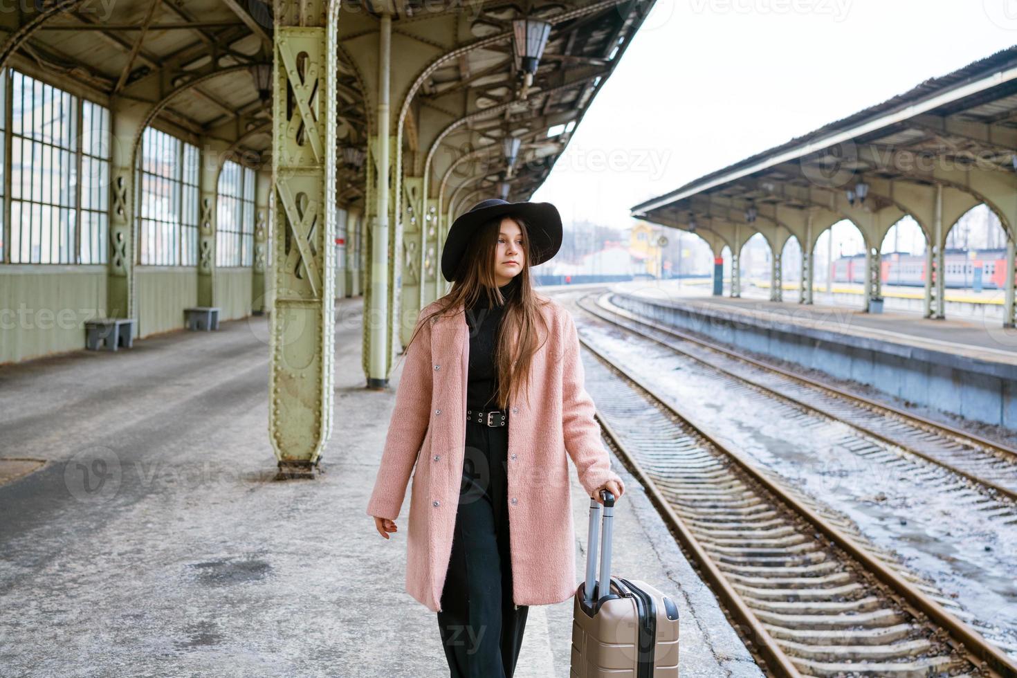 giovane donna in cappotto cammina con la valigia lungo la piattaforma vuota della ferrovia foto