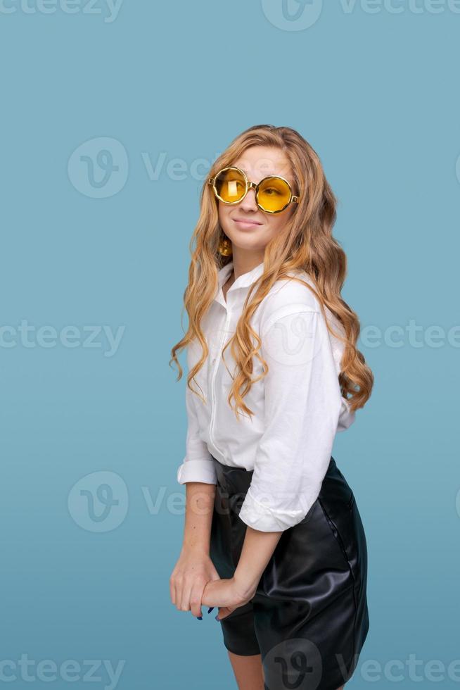 giovane donna allegra che indossa occhiali gialli sorridendo alla telecamera contro il blu foto