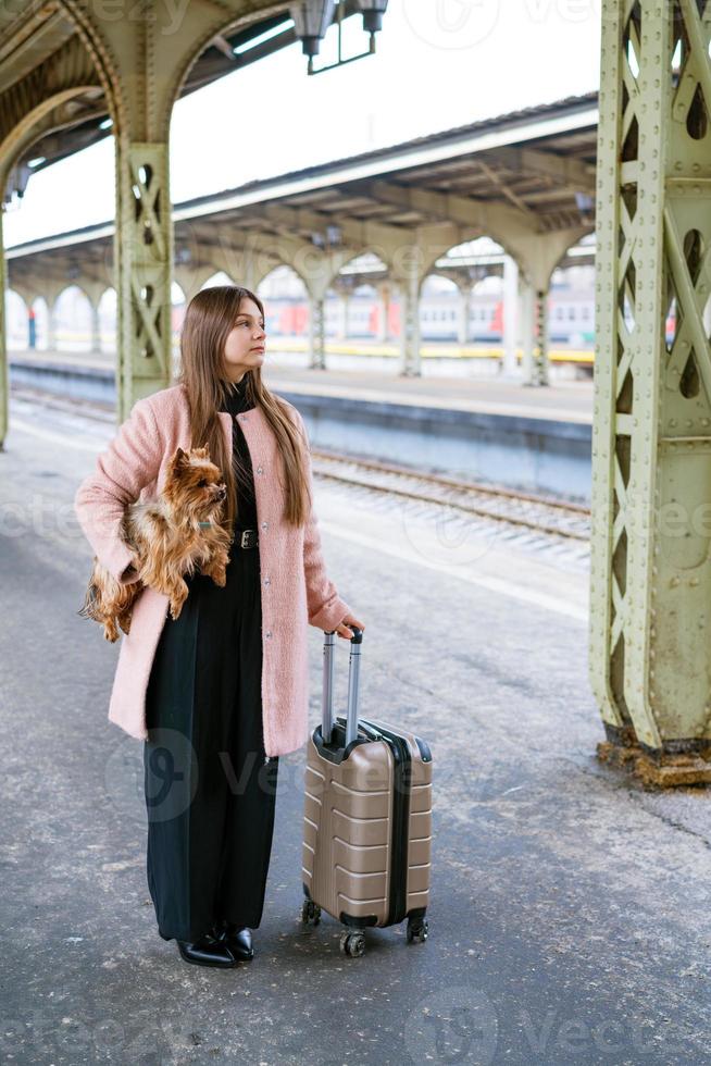il turista del viaggiatore della donna cammina con i bagagli e il cane alla stazione ferroviaria in rosa foto
