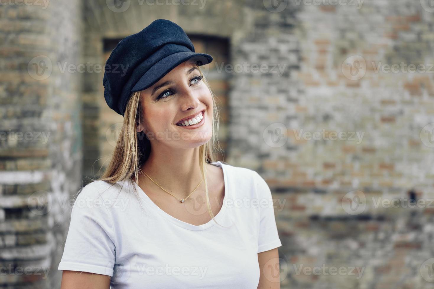 giovane donna bionda che indossa il cappello sorridente vicino a un muro di mattoni. foto