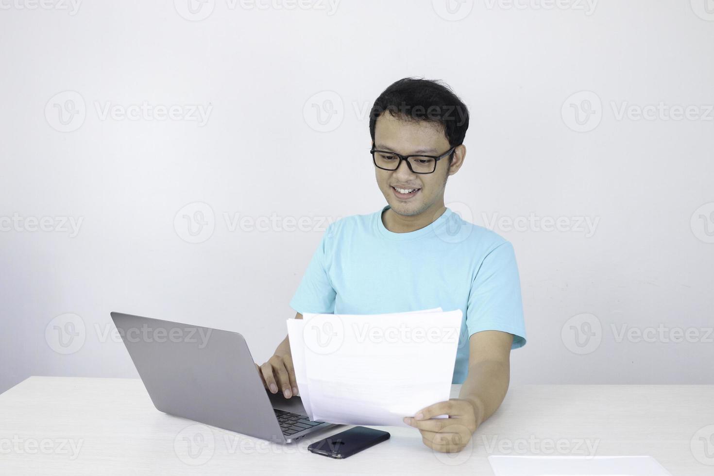 il giovane asiatico è sorridente e felice quando lavora su un laptop e ha un documento a portata di mano. uomo indonesiano che indossa una camicia blu. foto