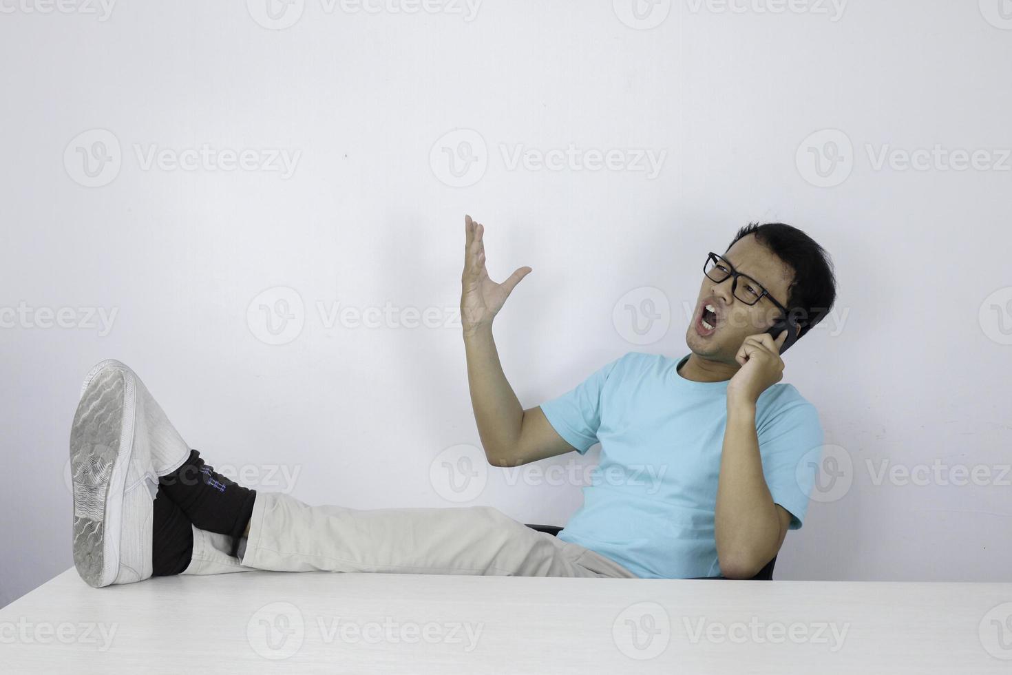 giovane uomo asiatico con la faccia scioccata sul telefono cellulare con la gamba sul tavolo. uomo indonesiano che indossa una camicia blu. foto