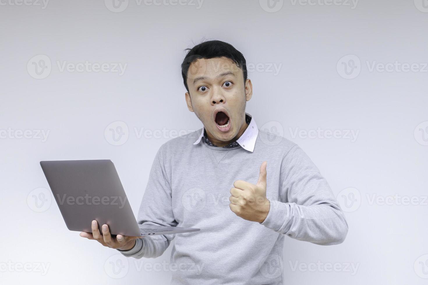 wow il viso del giovane asiatico ha scioccato ciò che vede nel laptop quando lavora su uno sfondo grigio isolato indossando una camicia grigia foto