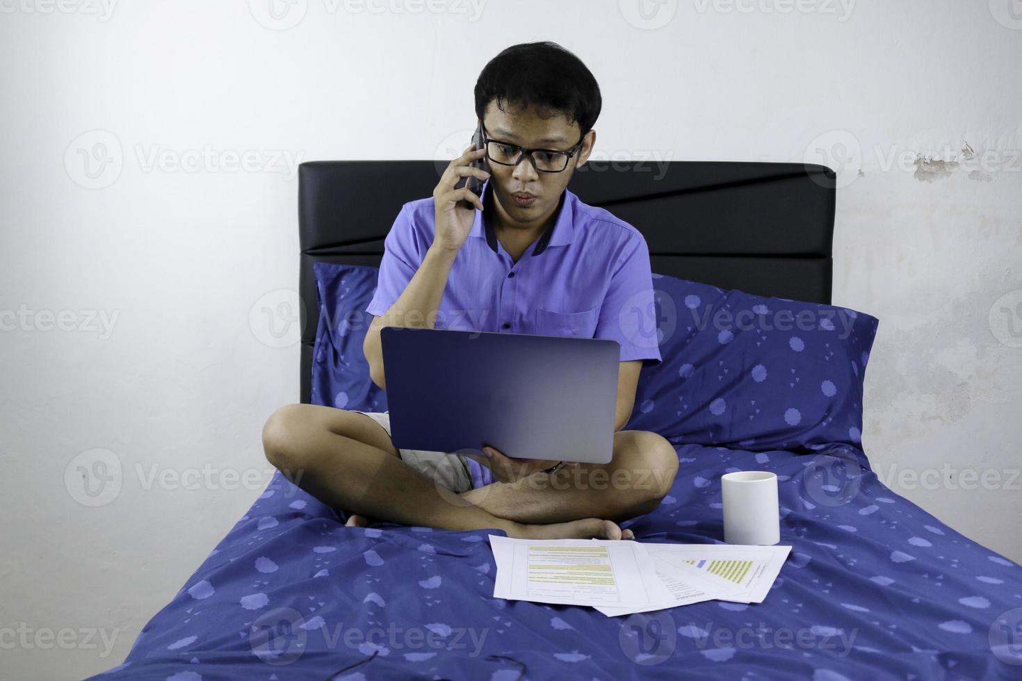 il giovane asiatico sta lavorando a casa con la chiamata in smartphone. un uomo seduto sul letto e al lavoro. concetto di lavoro da casa. foto