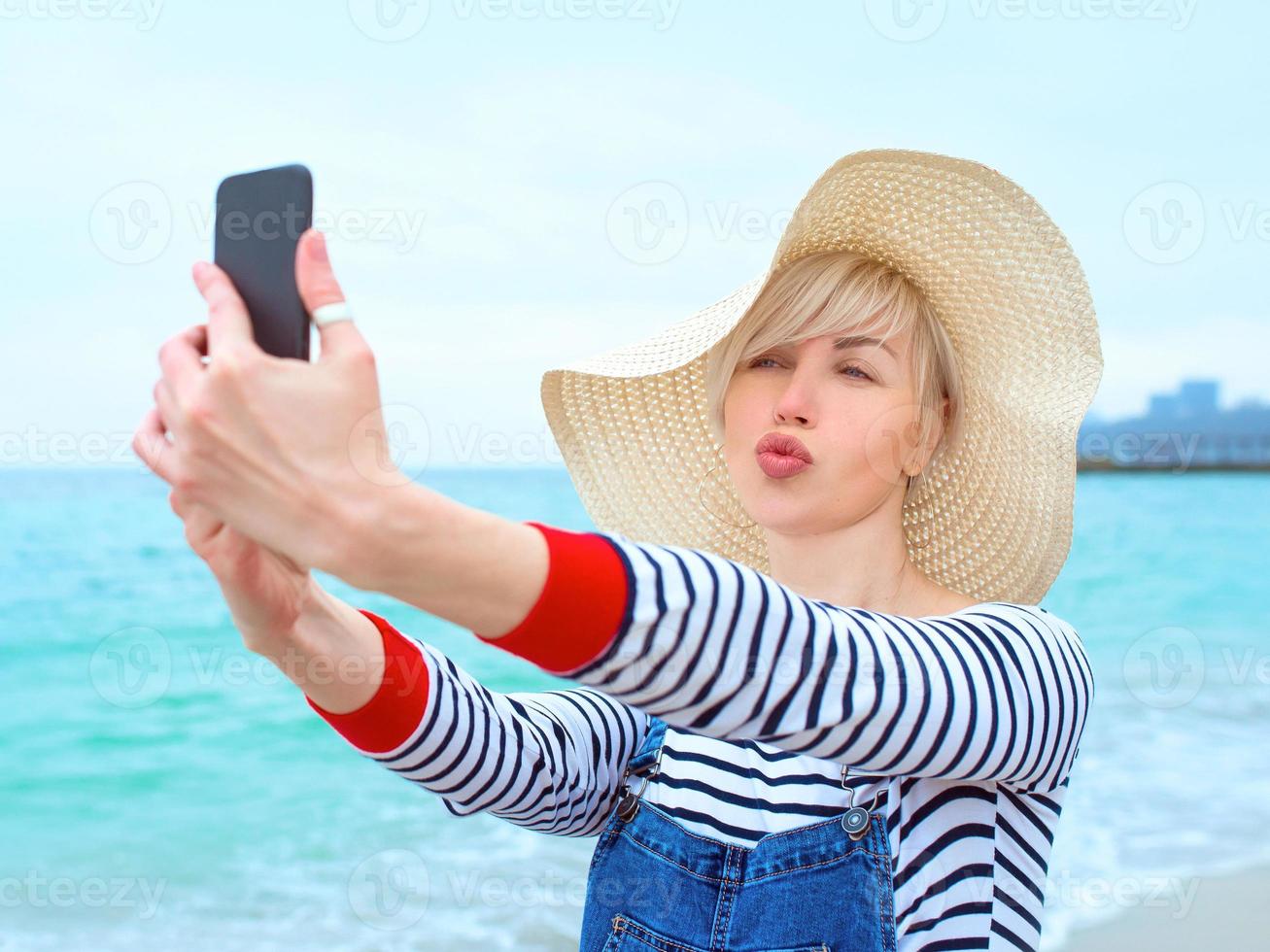 bella giovane donna caucasica bionda in vacanza con cappello di paglia, camicetta a righe e salopette di jeans che fanno selfie sullo smartphone sullo sfondo del mare blu incredibile foto