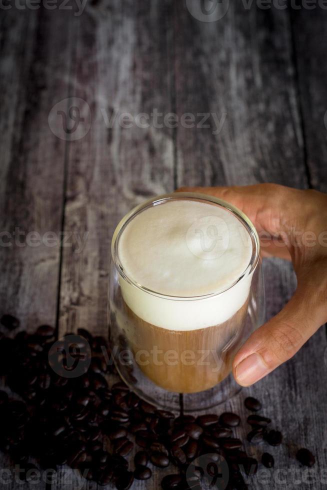 tempo del caffè e concetto di rinfresco. mano che tiene un bicchiere di caffè trasparente con schiuma di latte e chicco di caffè tostato su fondo di legno. foto