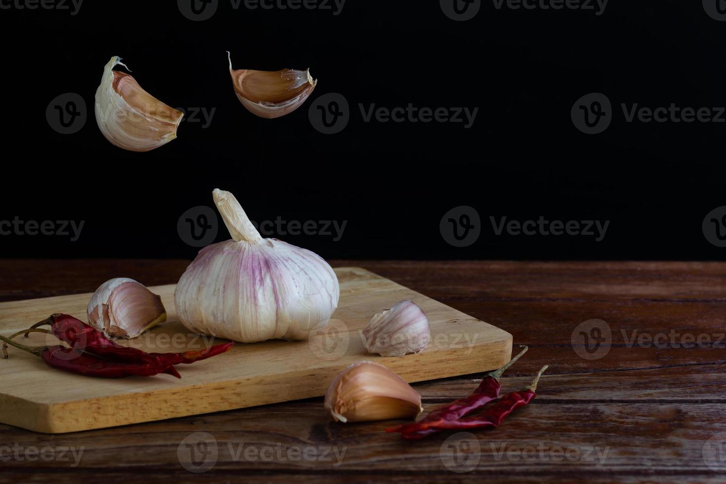 gruppo di aglio sul tagliere e alcuni spicchi d'aglio che galleggiano nell'aria e peperoncino rosso secco sul tavolo di legno con sfondo nero. copia spazio per il tuo testo. foto