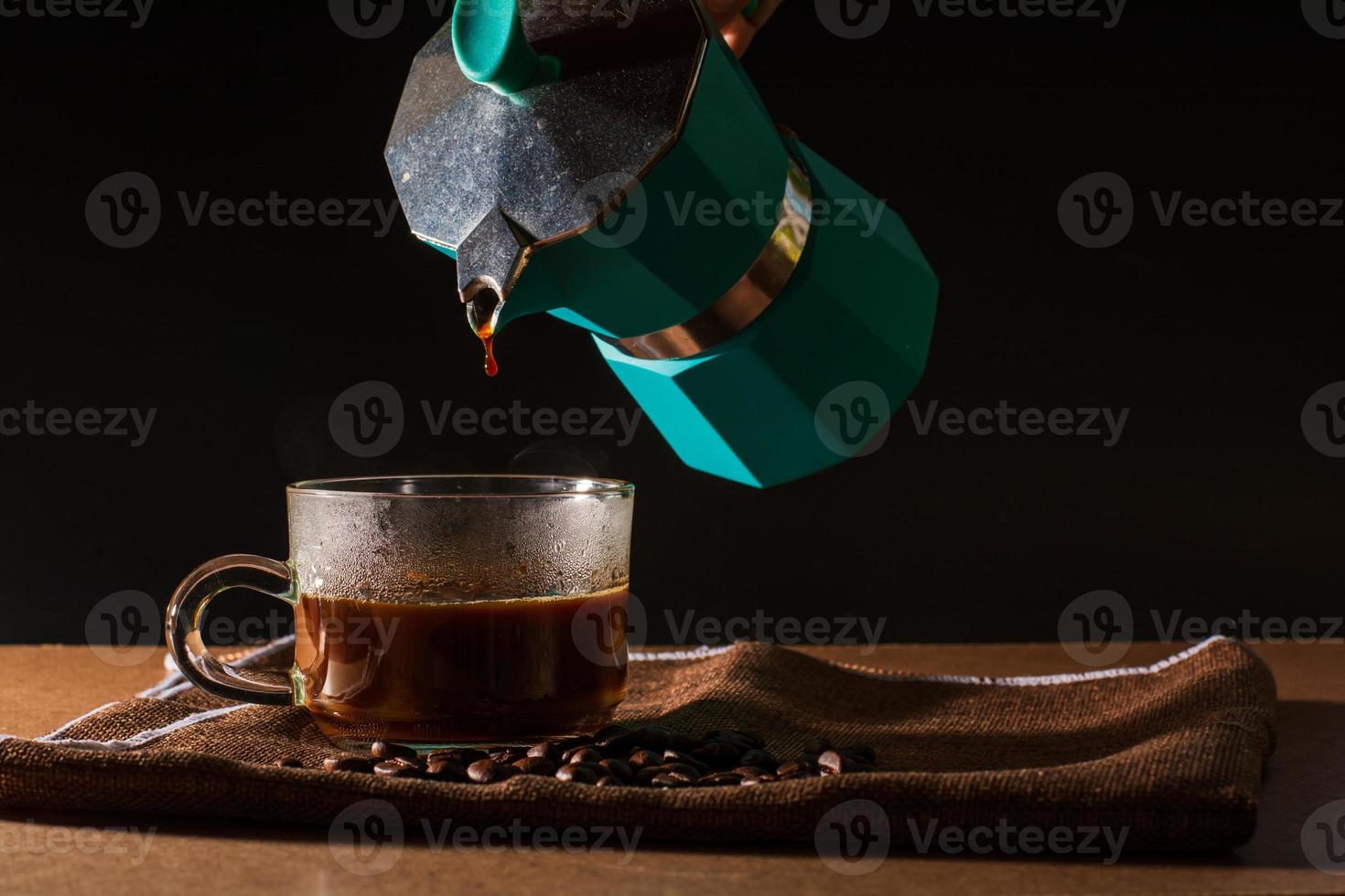 versare il caffè nero caldo dalla moka verde per pulire la tazza di caffè con fumo e chicchi di caffè su una tovaglia marrone e un tavolo di legno. beneficio del concetto di caffè. foto