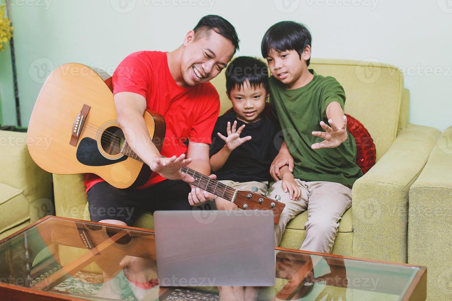 foto sfocata del padre asiatico con la chitarra che sorride insieme al laptop con suo figlio durante la videoconferenza