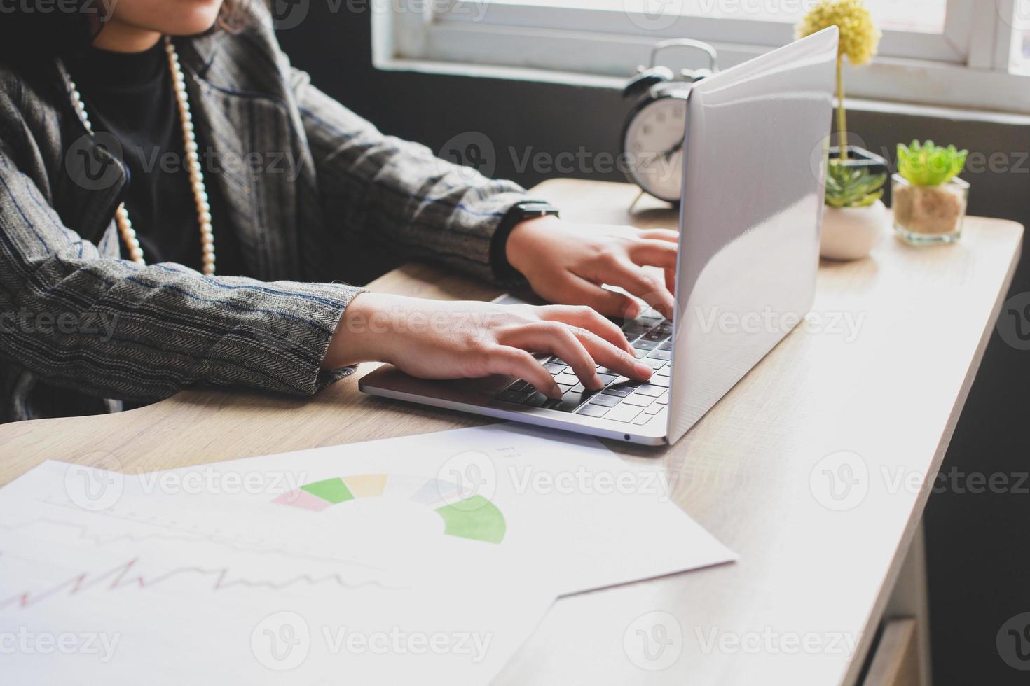 il colpo ritagliato della mano della donna d'affari sta digitando sulla tastiera del laptop con l'ufficio disordinato su una scrivania in legno foto