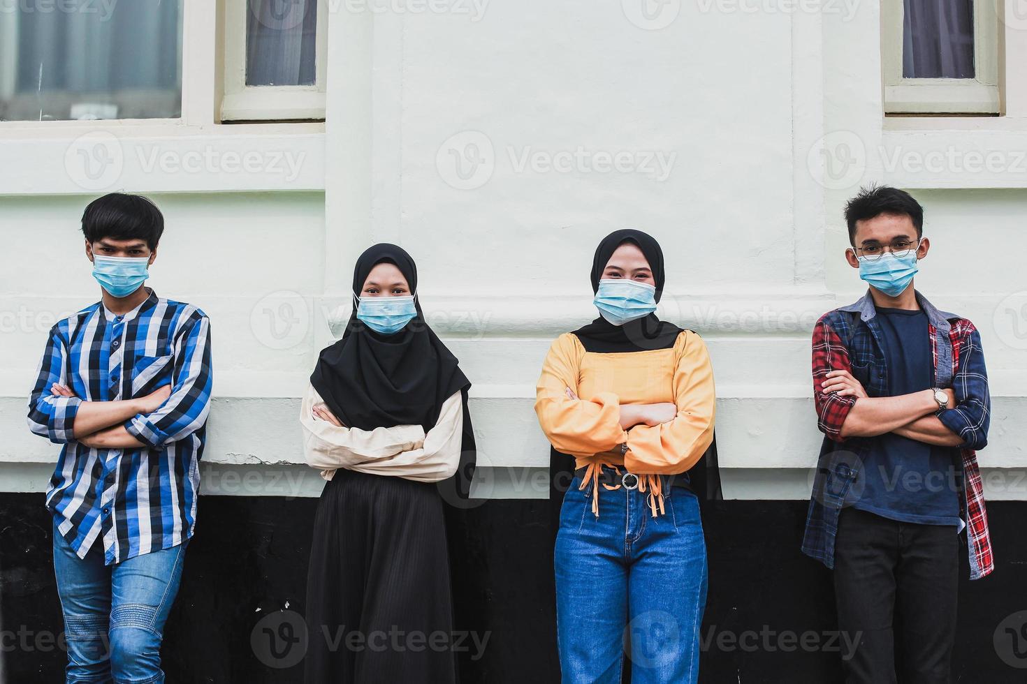 gruppo di adolescenti con la schiena appoggiata al muro e le braccia conserte che indossano maschere per proteggersi insieme dal coronavirus foto