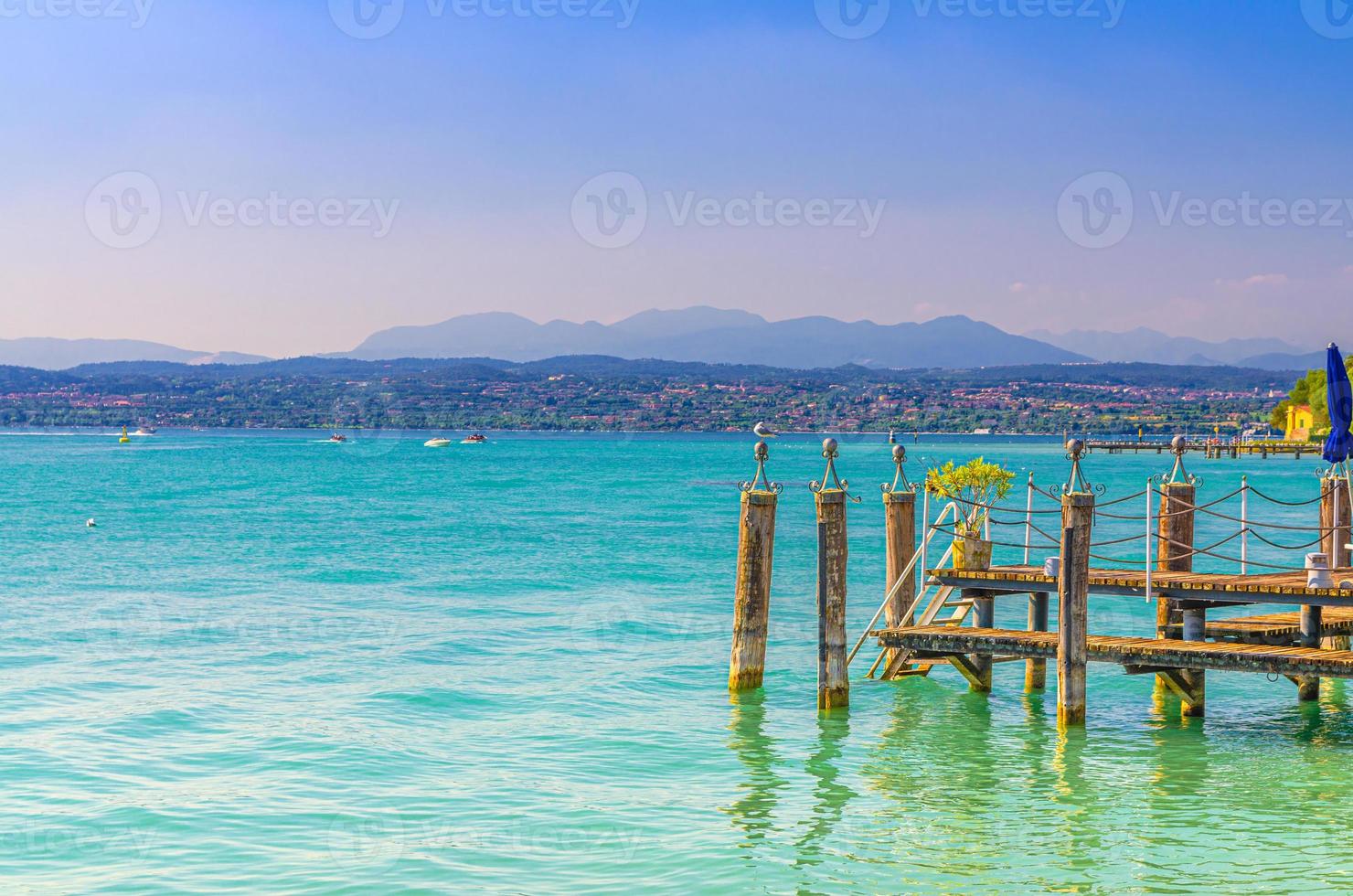 lago di garda con acque turchesi azzurre e molo in legno, costa con catena montuosa foto