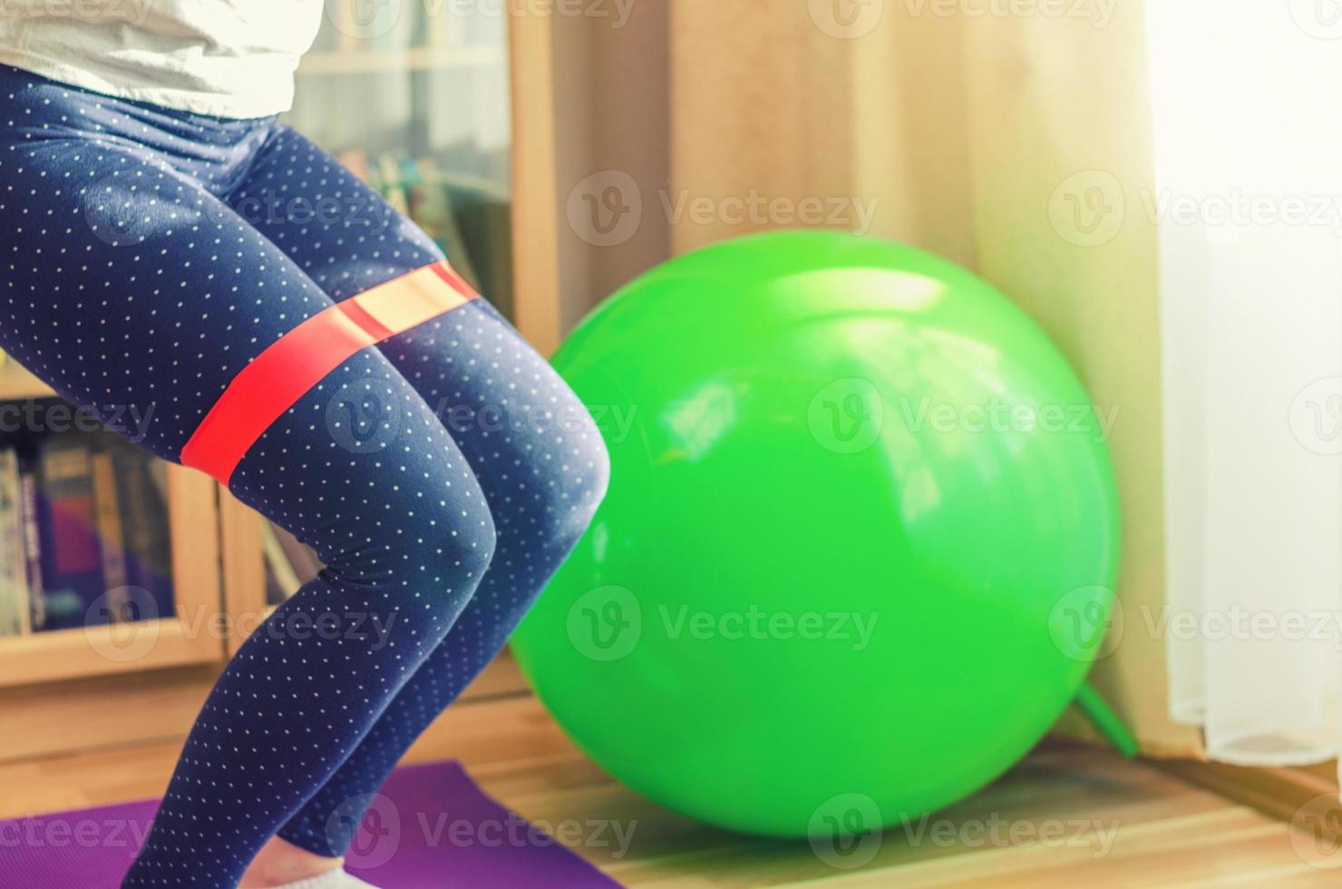 gambe di donna in leggings sportivi che fanno sit-up ginocchia squat con fascia di resistenza in gomma rossa durante l'allenamento a casa, palla fitness in gomma verde foto