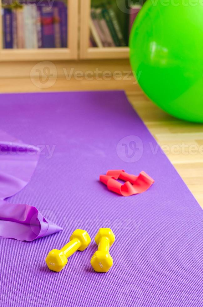 attrezzatura sportiva sul pavimento del soggiorno, tappetino da yoga viola, manubri gialli, fascia di resistenza in gomma rossa e palla aerobica fitness in gomma verde foto