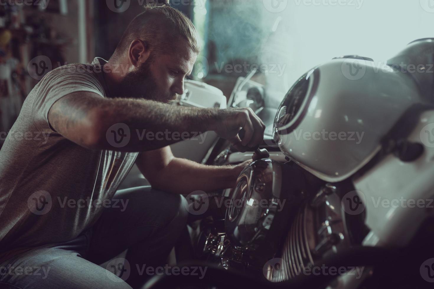 bell'uomo barbuto che ripara la sua moto in garage. un uomo che indossa jeans e una maglietta foto