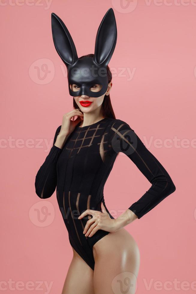 donna sexy che indossa un coniglietto di pasqua con maschera nera in piedi su uno sfondo rosa e sembra molto sensuale foto