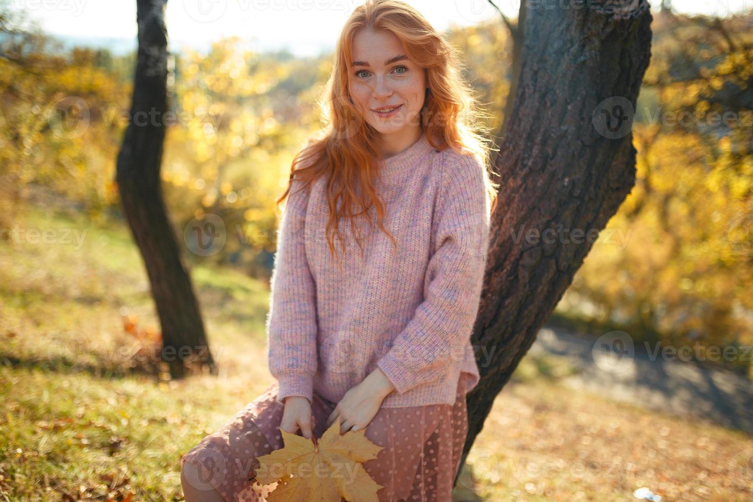 ritratti di un'affascinante ragazza dai capelli rossi con un viso carino. ragazza in posa nel parco autunnale con un maglione e una gonna color corallo. nelle mani di una ragazza una foglia gialla foto