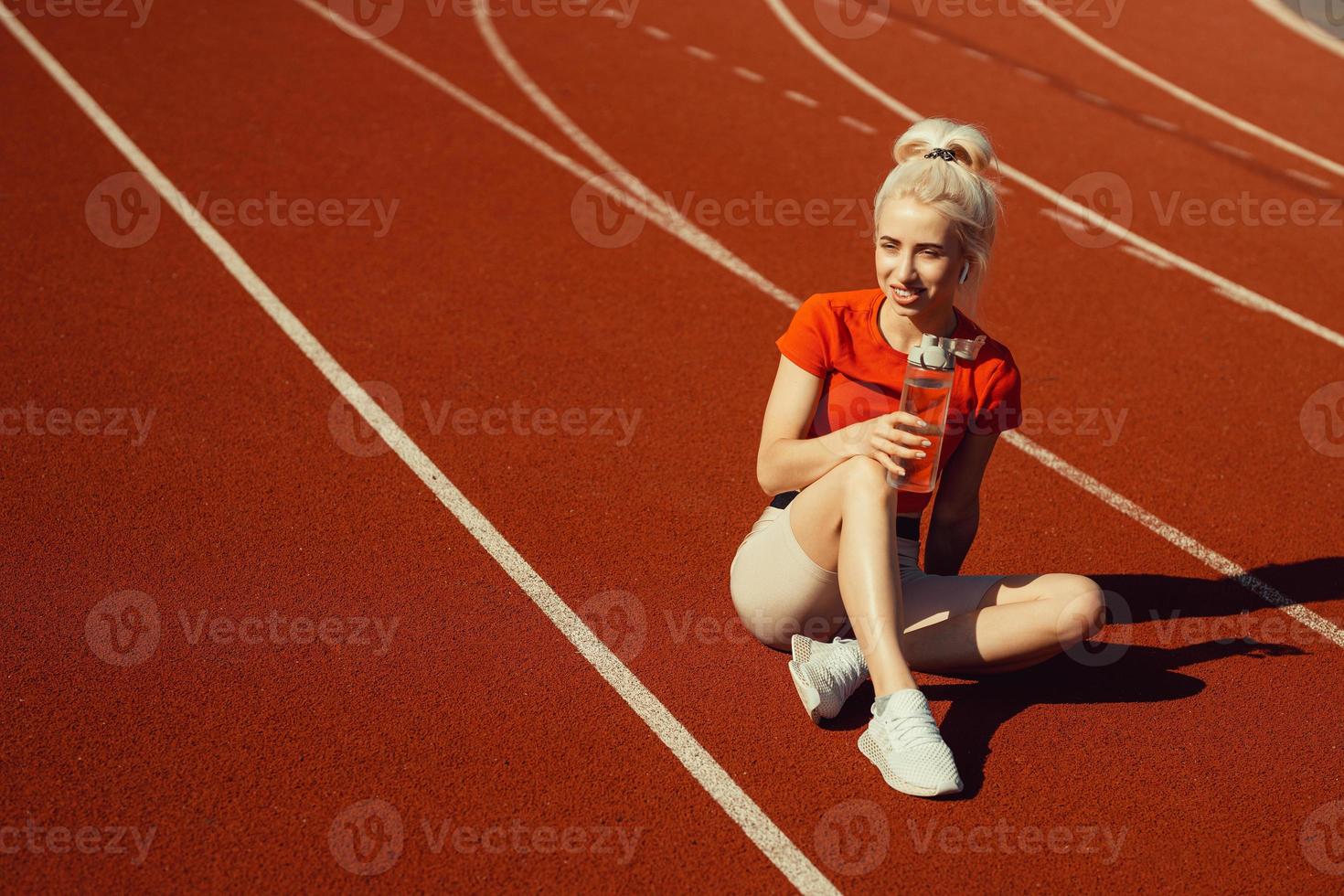 giovane bella bionda si siede su una pista da jogging con una bottiglia d'acqua in mano foto