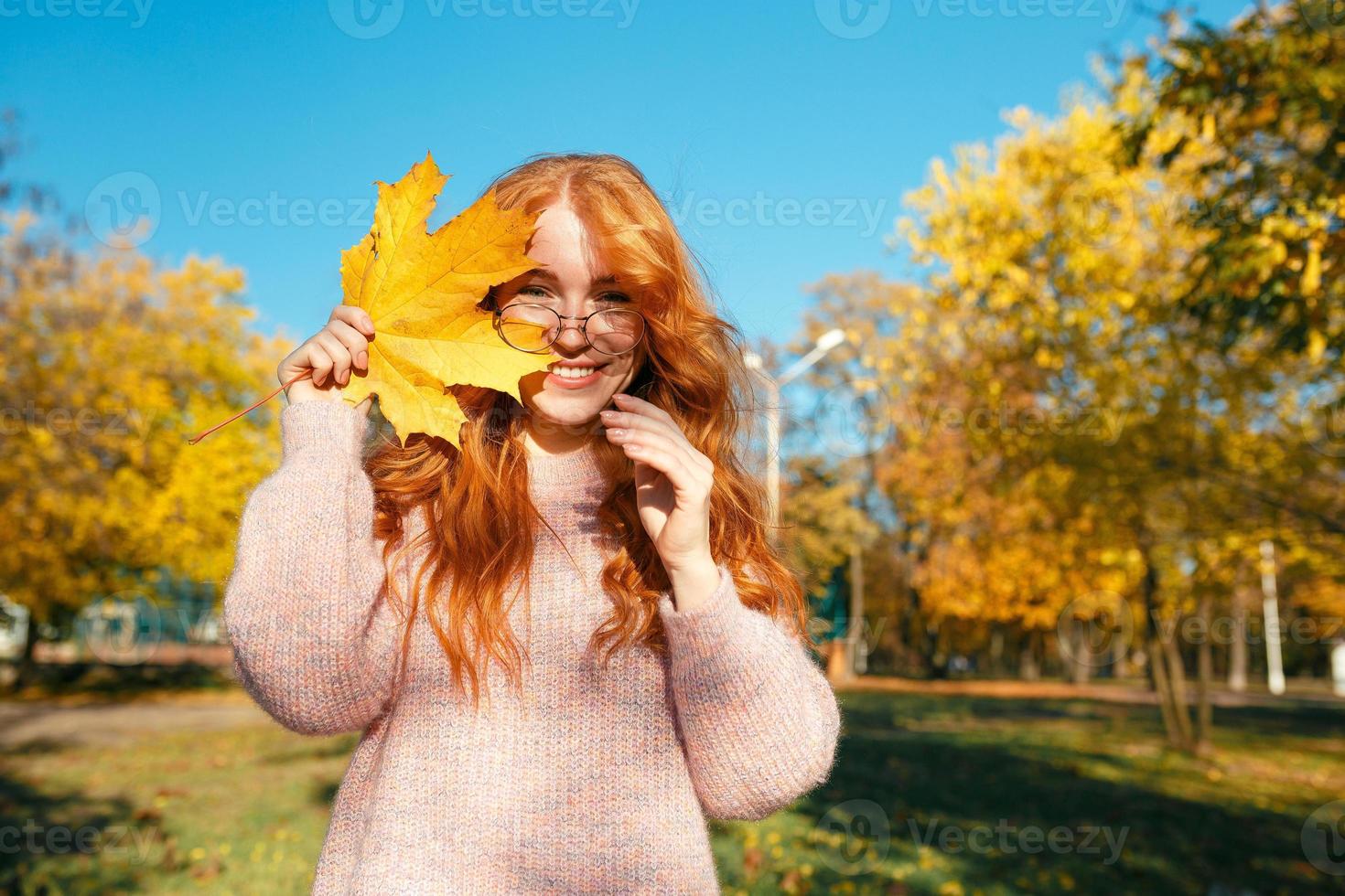 ritratti di un'affascinante ragazza dai capelli rossi con un viso carino. ragazza in posa nel parco autunnale con un maglione e una gonna color corallo. nelle mani di una ragazza una foglia gialla foto