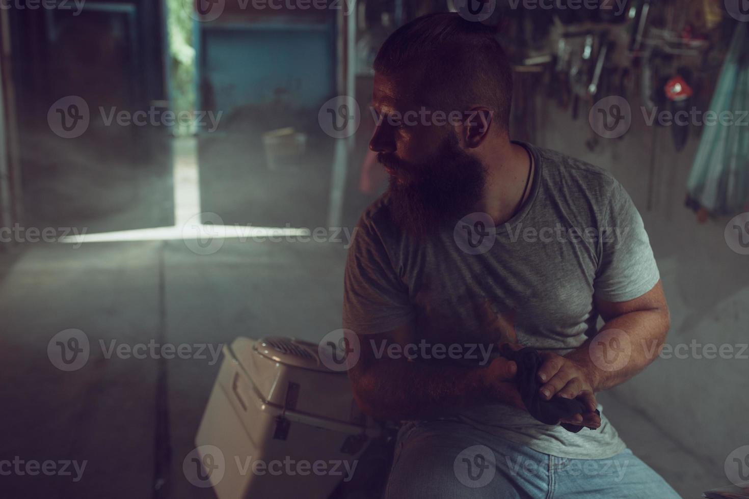 bell'uomo brutale con la barba seduto su una motocicletta nel suo garage, asciugandosi le mani e guardando di lato foto