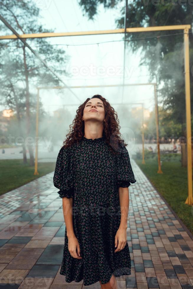 la simpatica ragazza riccia dai capelli rossi si rallegra sotto il nebulizzatore d'acqua nel centro della città foto