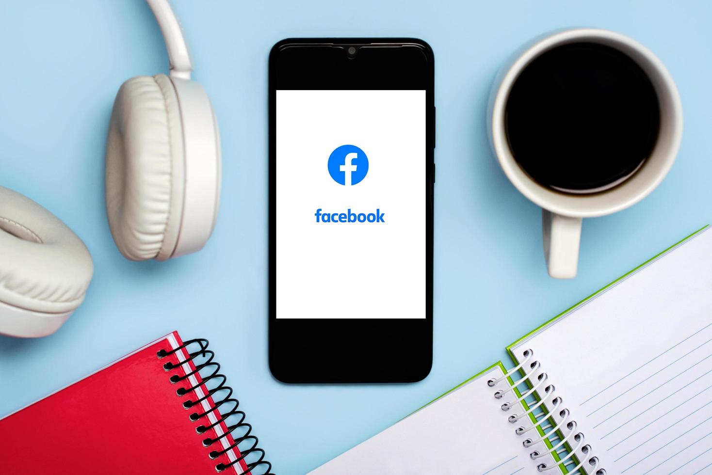 logo facebook sullo schermo bianco dello smartphone con notebook, cuffie e tazza di caffè foto