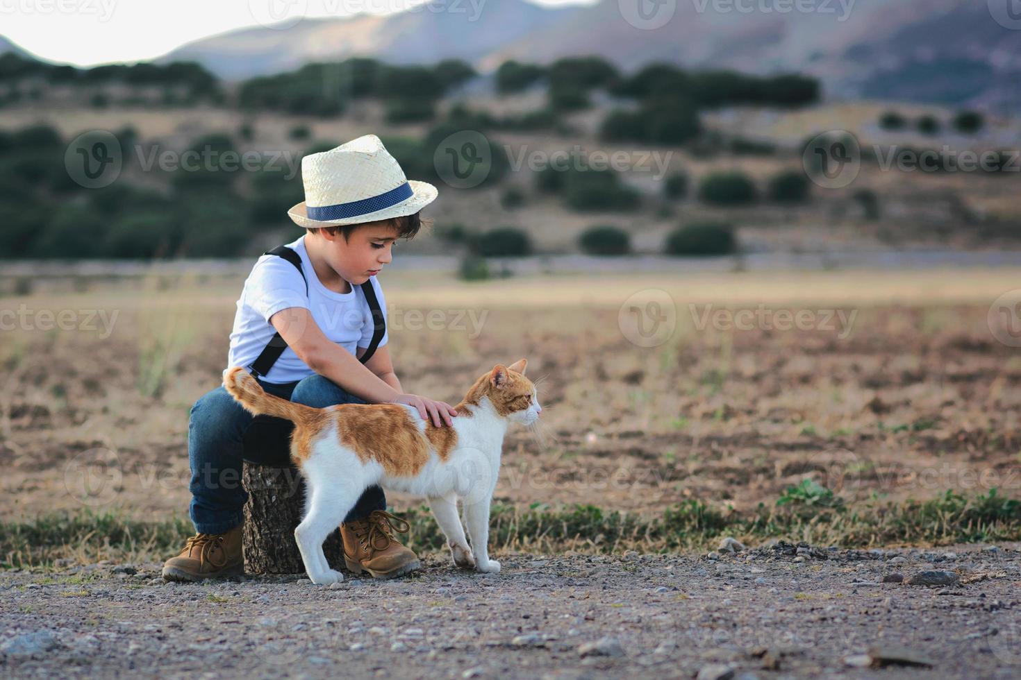 bambino con maglietta bianca e cappello seduto su un tronco di legno accarezza il suo gatto foto