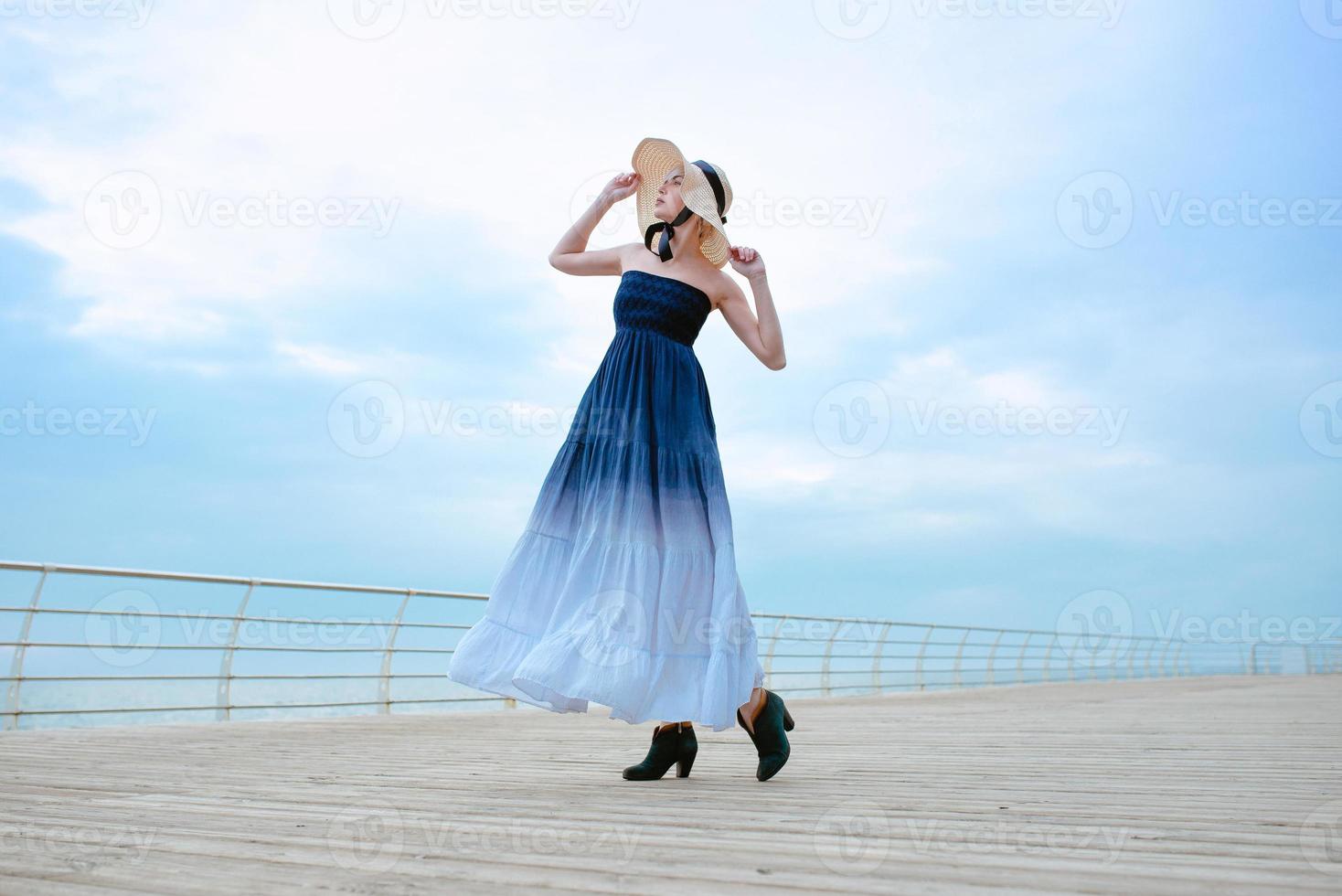 caucasica bella bionda donna triste in abito blu e cappello di paglia in piedi vicino al mare foto