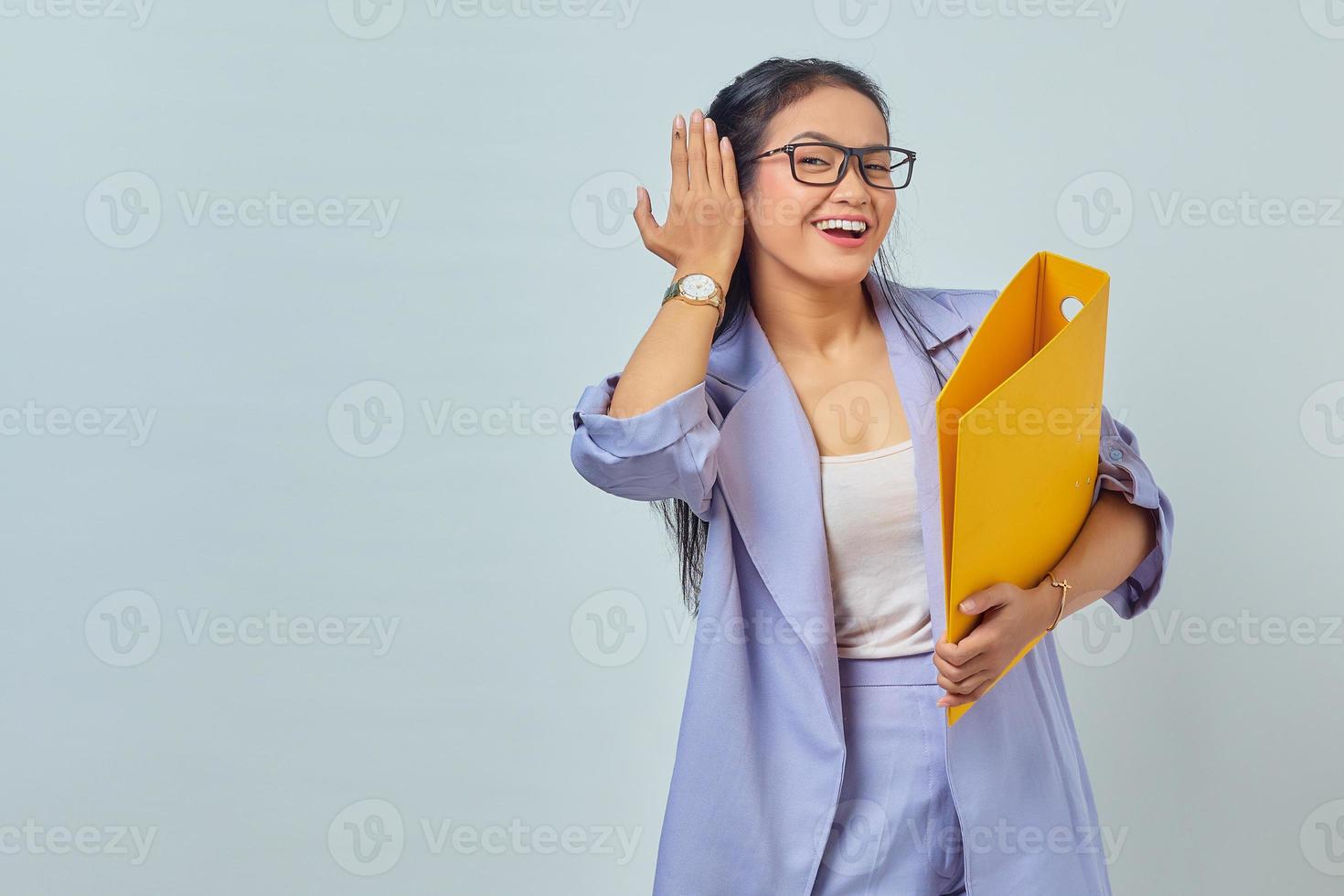il ritratto di una giovane donna d'affari asiatica seria tiene la mano vicino all'orecchio cerca di ascoltare la conversazione privata ascolta i pettegolezzi mentre tiene la cartella gialla isolata su sfondo viola foto