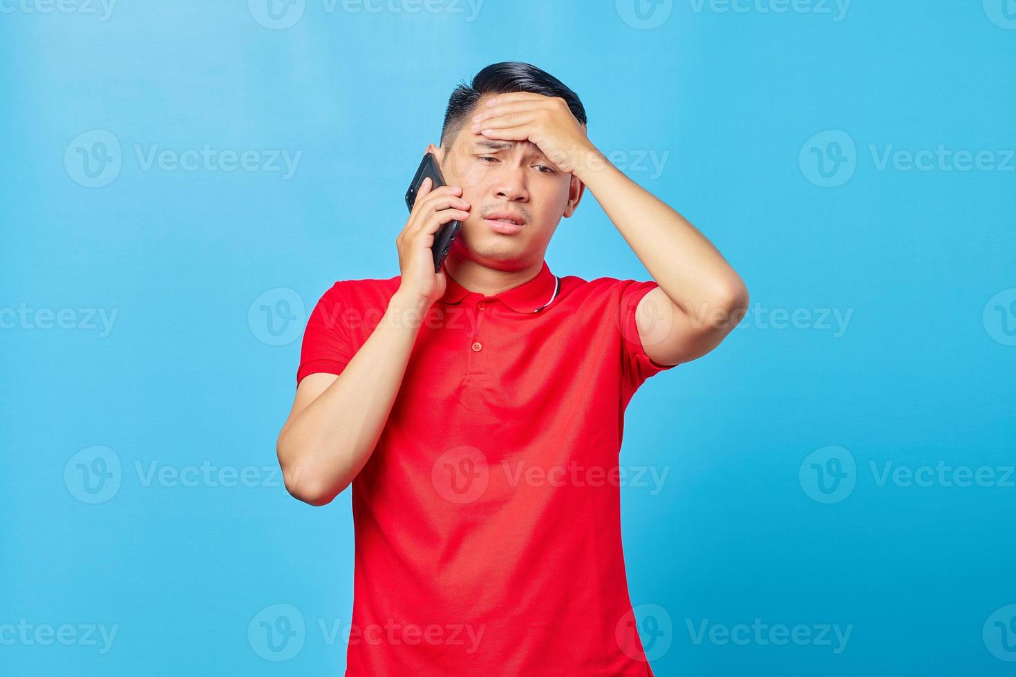 ritratto di un uomo asiatico vertiginoso che parla sullo smartphone e tiene la testa con il palmo isolato su sfondo blu foto