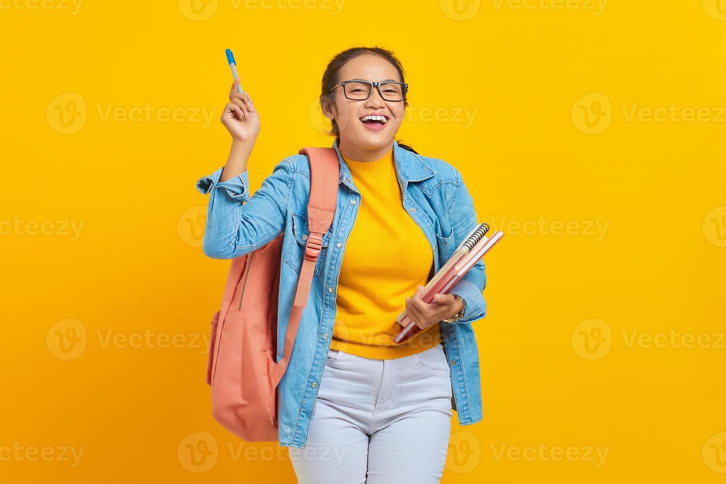 ritratto di giovane studentessa asiatica allegra in abiti casual con libro in possesso di zaino e che punta allo spazio della copia con penna isolato su sfondo giallo. istruzione nel concetto di università universitaria foto