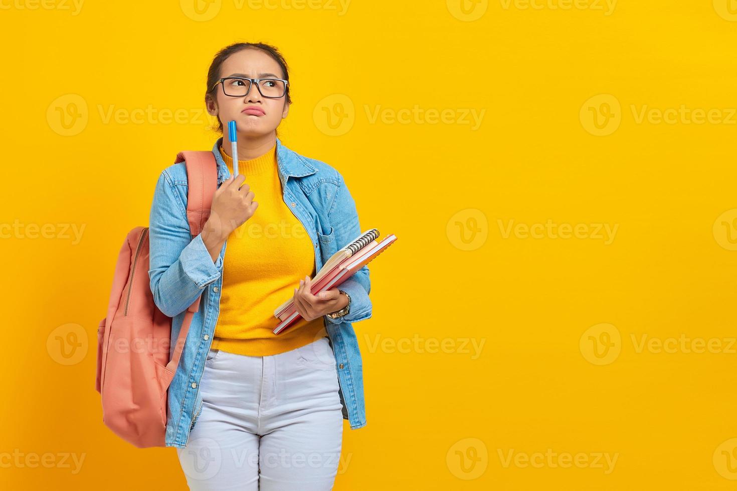 Ritratto di giovane studentessa asiatica seria in abiti casual con zaino che tiene libro e penna, pensando alla domanda isolata su sfondo giallo. istruzione nel concetto di università universitaria foto