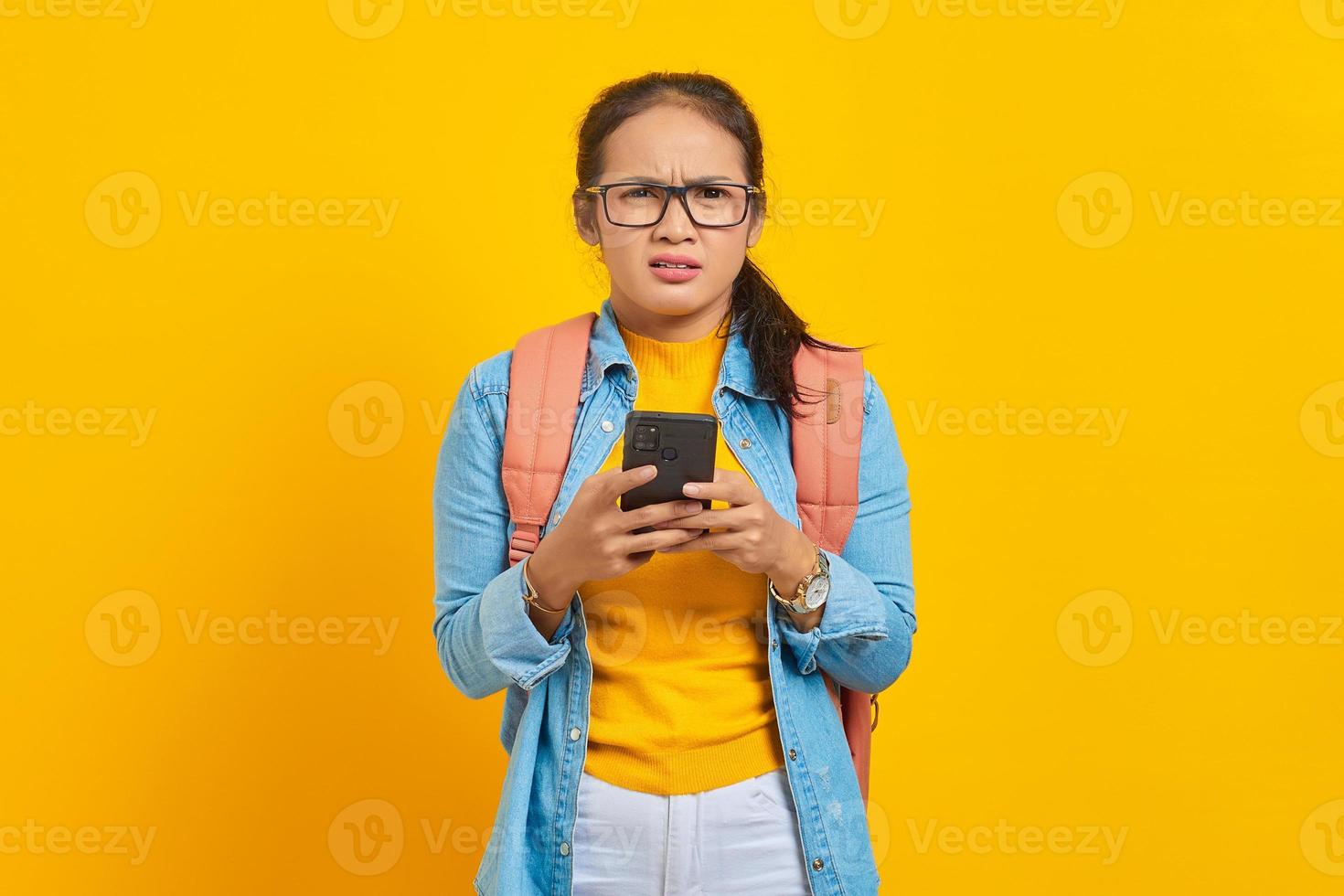 ritratto di giovane studentessa asiatica infastidita in abiti casual con zaino utilizzando il telefono cellulare e guardando la fotocamera isolata su sfondo giallo. istruzione nel concetto di università universitaria foto