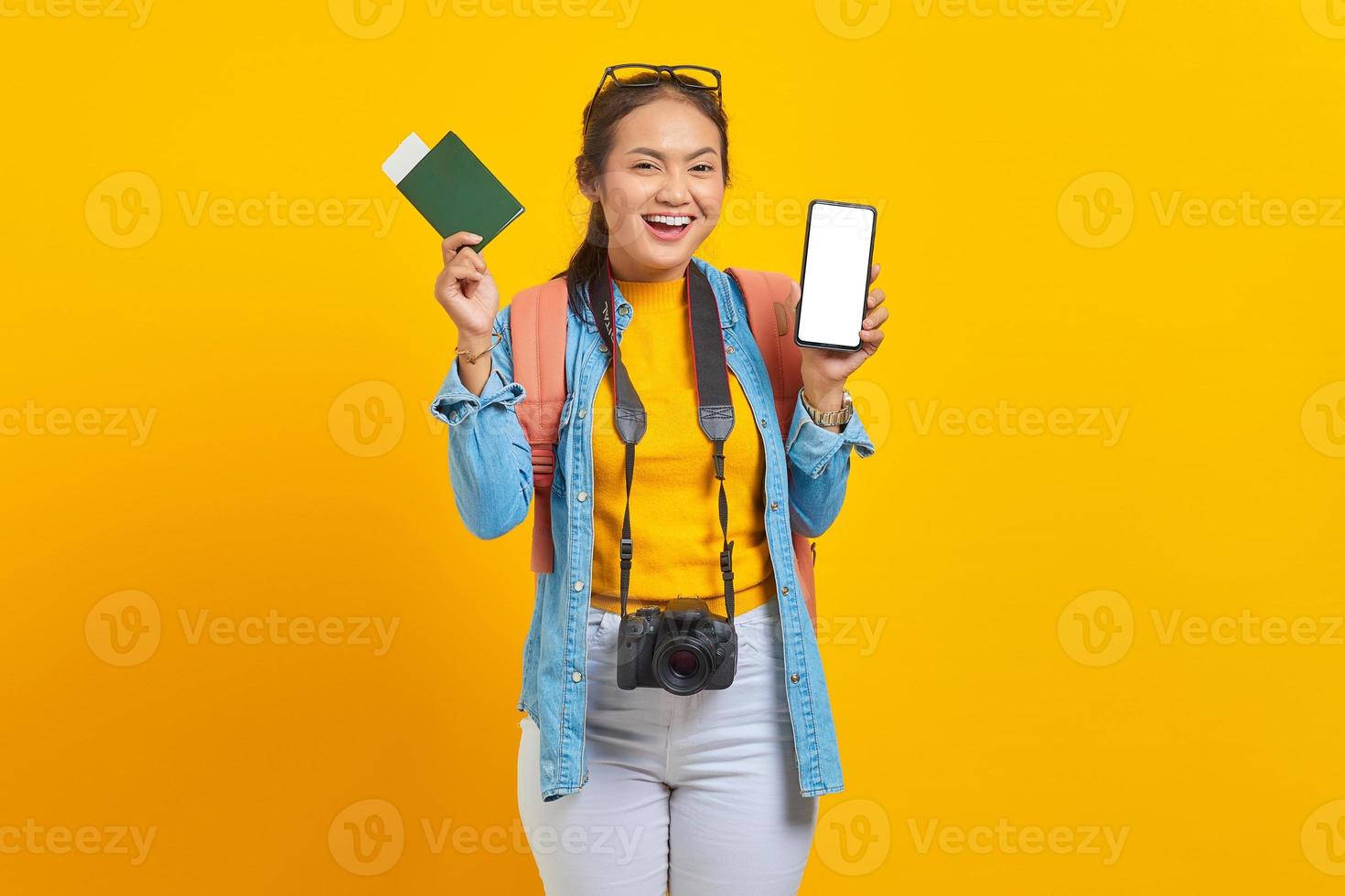ritratto di allegro giovane viaggiatore asiatico che mostra il biglietto della carta d'imbarco del passaporto e il telefono cellulare con schermo vuoto isolato su sfondo giallo. passeggeri che viaggiano nei fine settimana. concetto di viaggio aereo foto