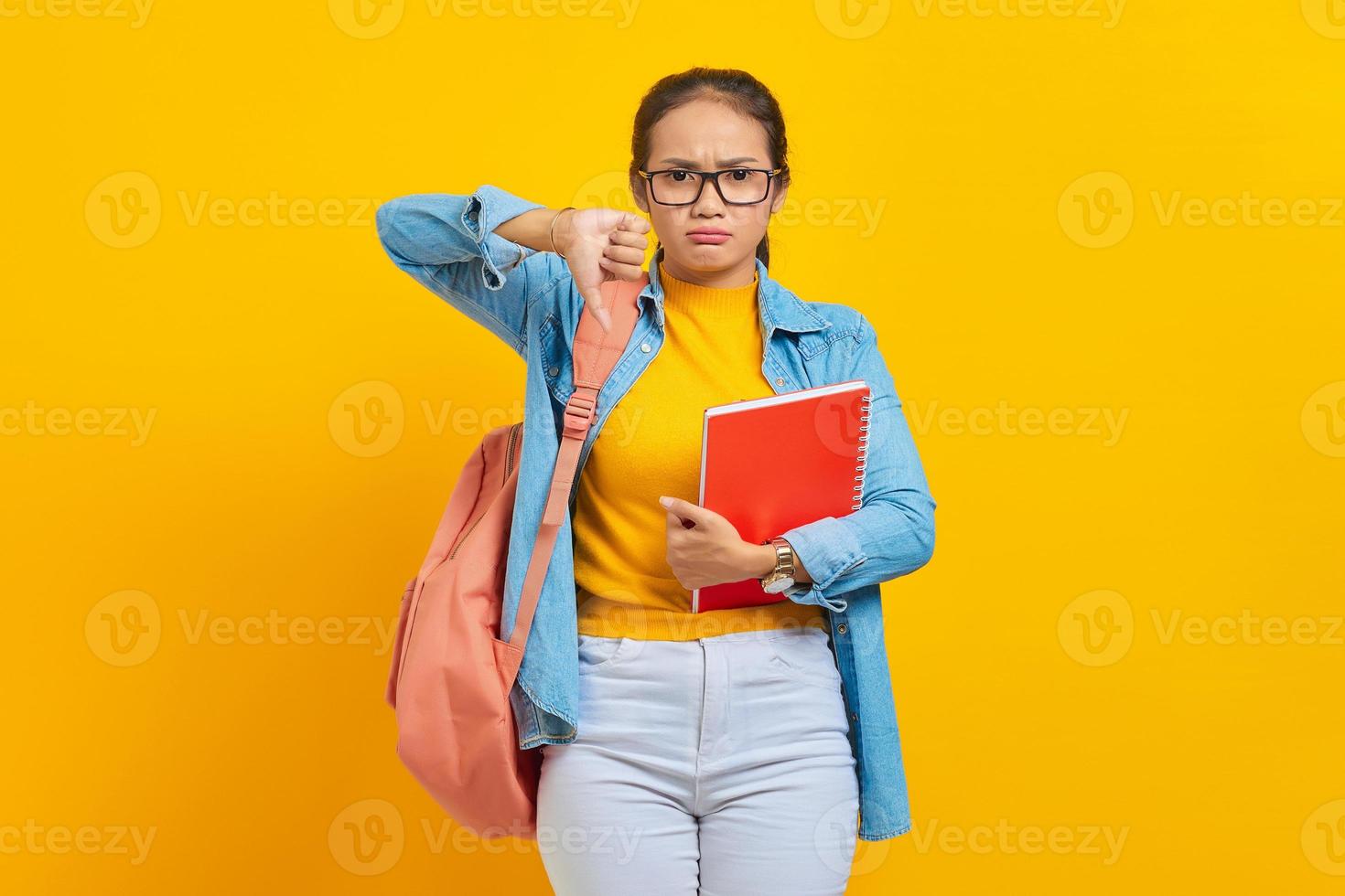 dispiaciuto preoccupato giovane studentessa asiatica in abiti denim con zaino in possesso di taccuino, mostrando il pollice verso il basso mentre si guarda da parte isolato su sfondo giallo foto