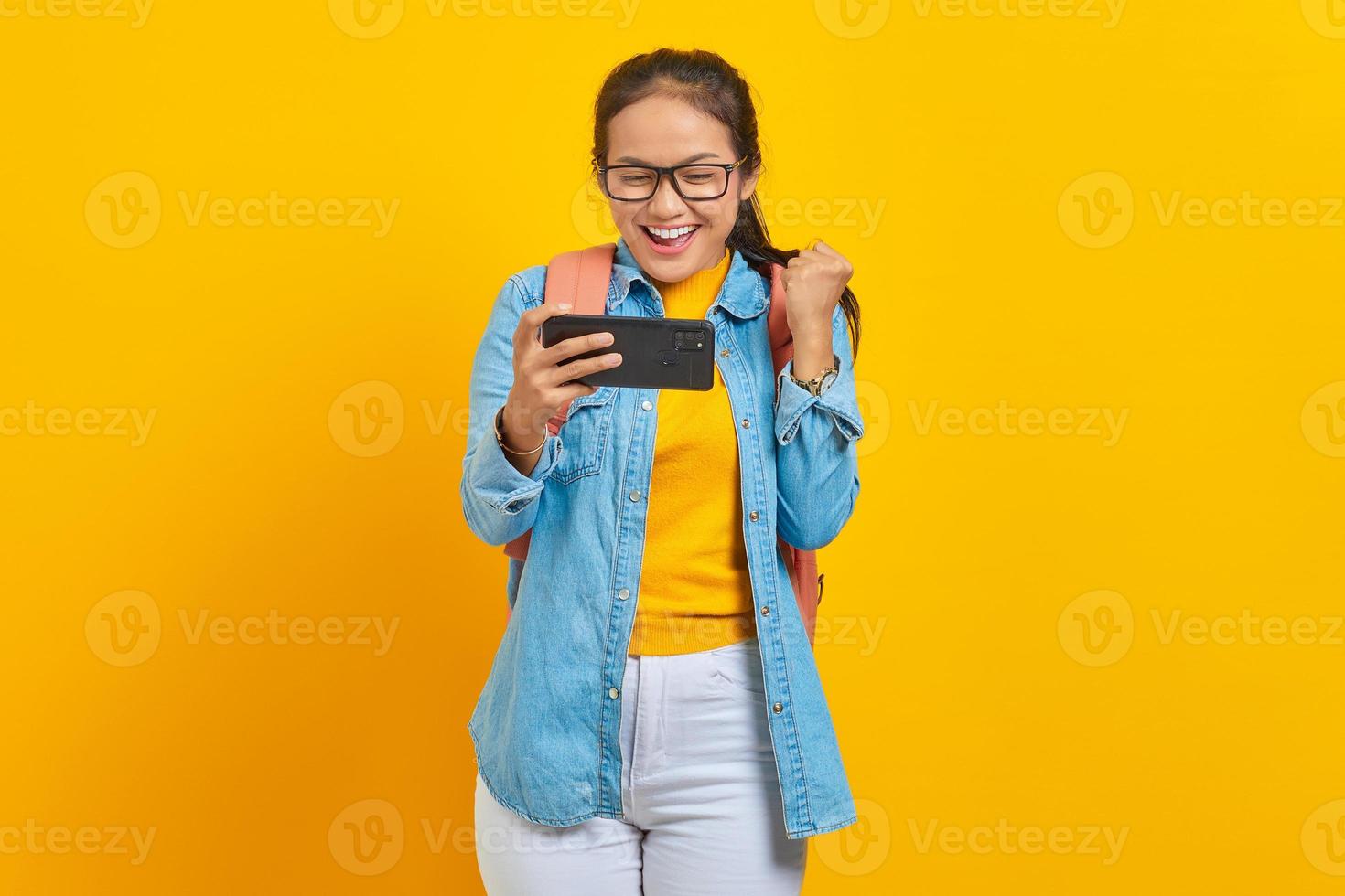 ritratto di giovane studentessa asiatica allegra in abiti casual con zaino che gioca al videogioco sul telefono cellulare e celebra il successo isolato su sfondo giallo foto