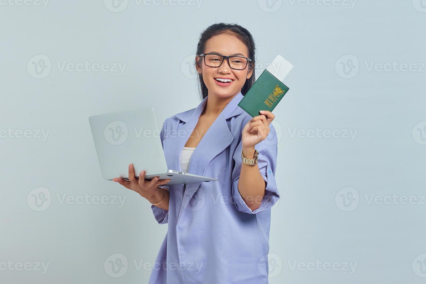 ritratto di giovane donna asiatica in piedi utilizzando il computer portatile e tenendo il libro del passaporto con espressione allegra isolato su sfondo bianco foto