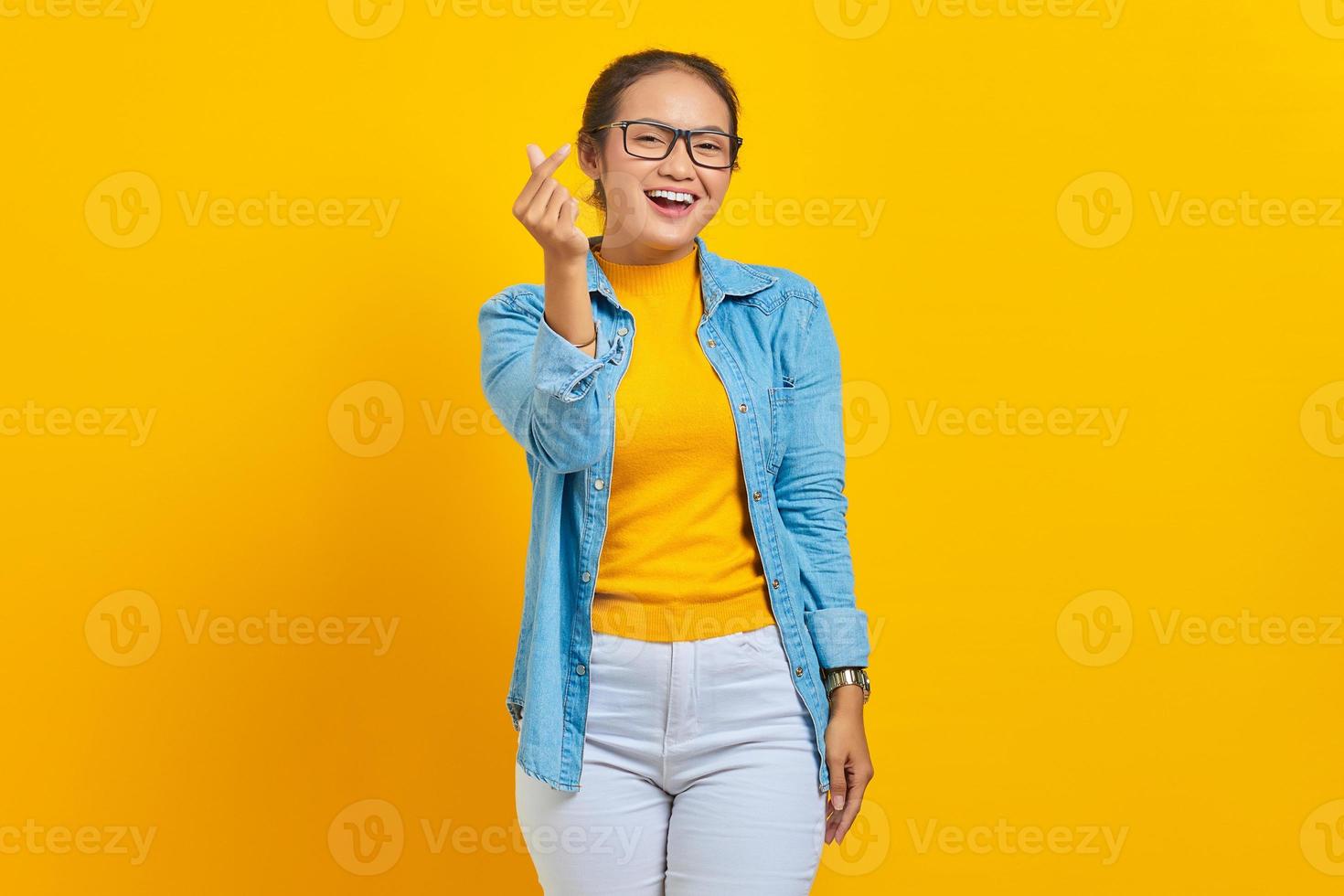 Ritratto di giovane studentessa asiatica sorridente in abiti in denim che mostra il cuore coreano con due dita incrociate isolato su sfondo giallo. persone emozioni sincere concetto di stile di vita foto