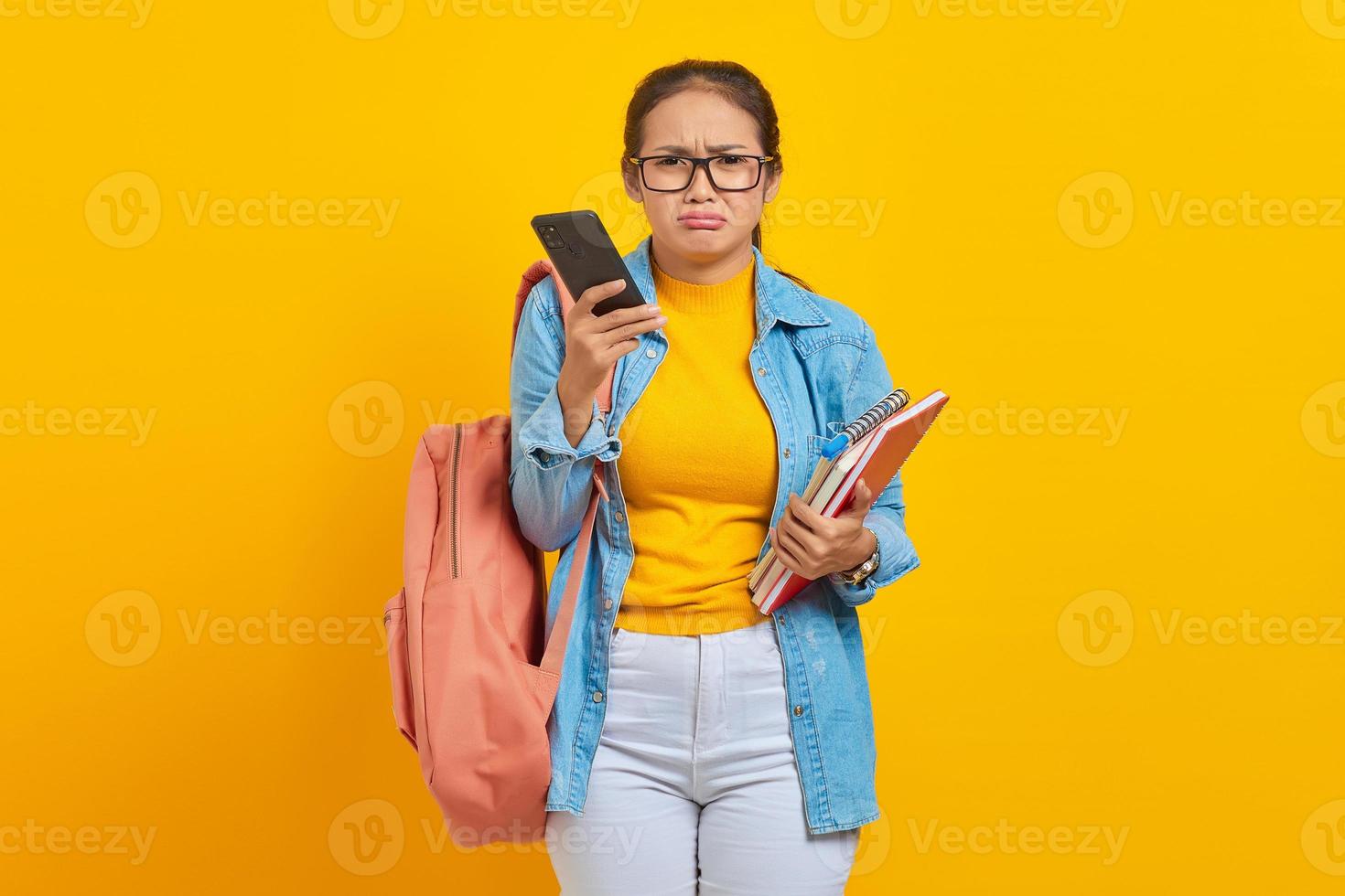 Ritratto di giovane studentessa asiatica triste in abiti denim con zaino utilizzando il telefono cellulare e tenendo il libro isolato su sfondo giallo foto