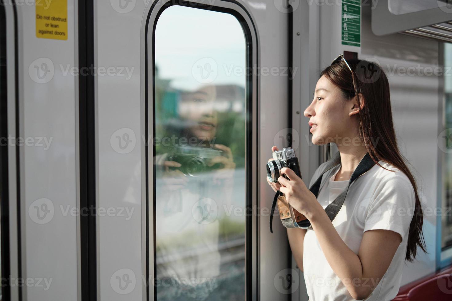 una bella turista asiatica con la macchina fotografica nella cabina passeggeri, che viaggia in treno sopraelevato, scatta foto istantanee durante il trasporto in vista urbana, stile di vita cittadino in treno, felice vacanza di viaggio.