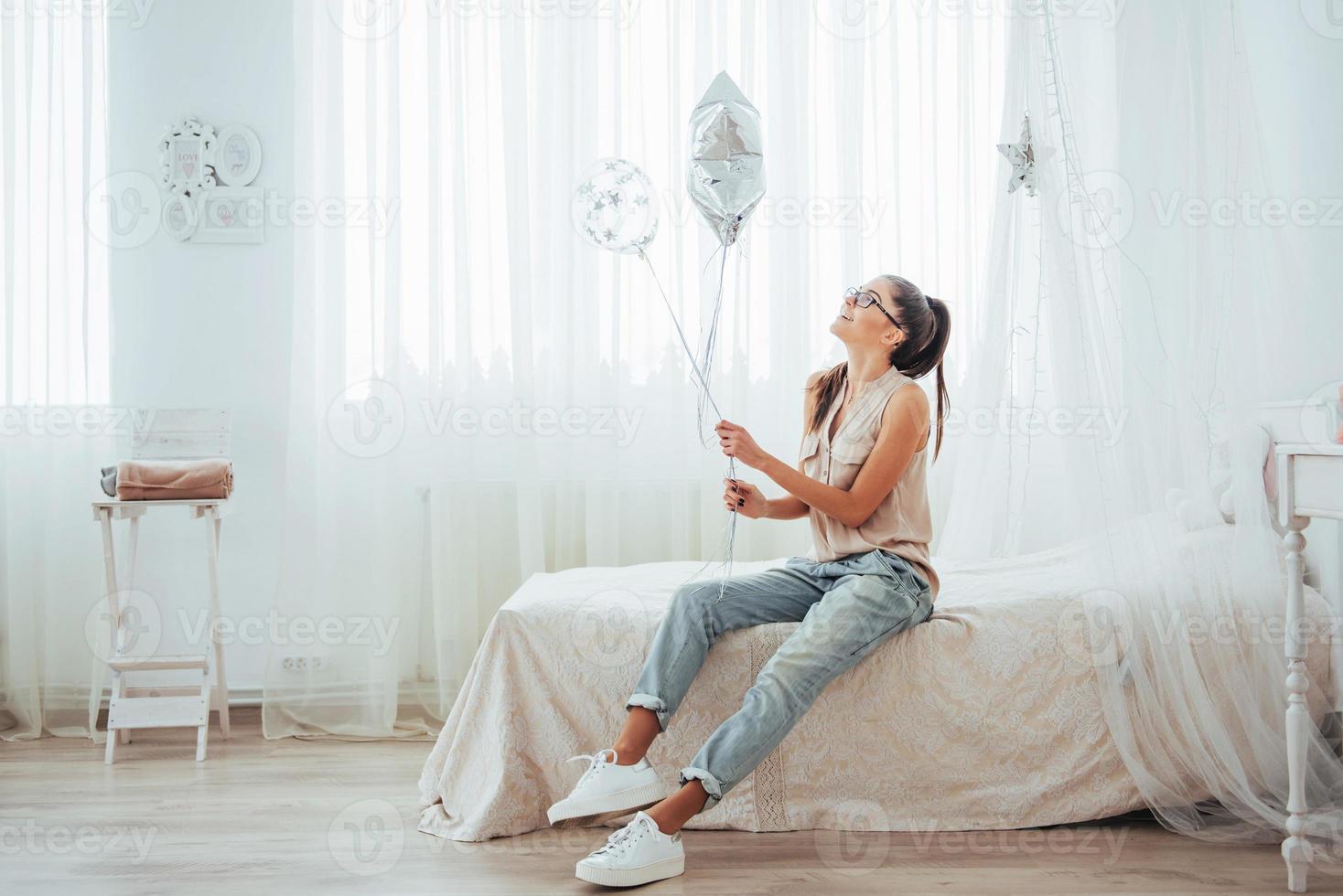 primo piano ragazza bruna carina in studio, sorridente ampiamente e giocando con palloncini trasparenti e d'argento. porta gli occhiali e i capelli intrecciati. foto