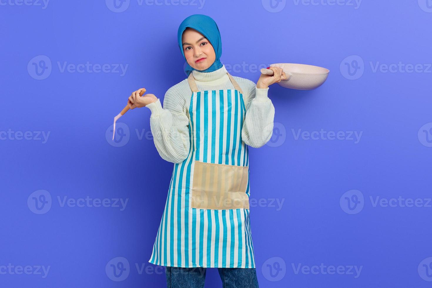 ritratto di giovane casalinga musulmana asiatica confusa che indossa un grembiule che tiene una spatola e una padella mentre fa i lavori domestici isolati su sfondo viola. concetto di pulizia foto