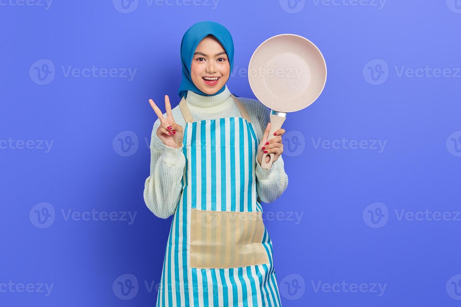 allegra bella donna asiatica in grembiule e hijab che tiene padella e mostra il segno di pace con le dita isolate su sfondo viola. concetto di pulizia foto