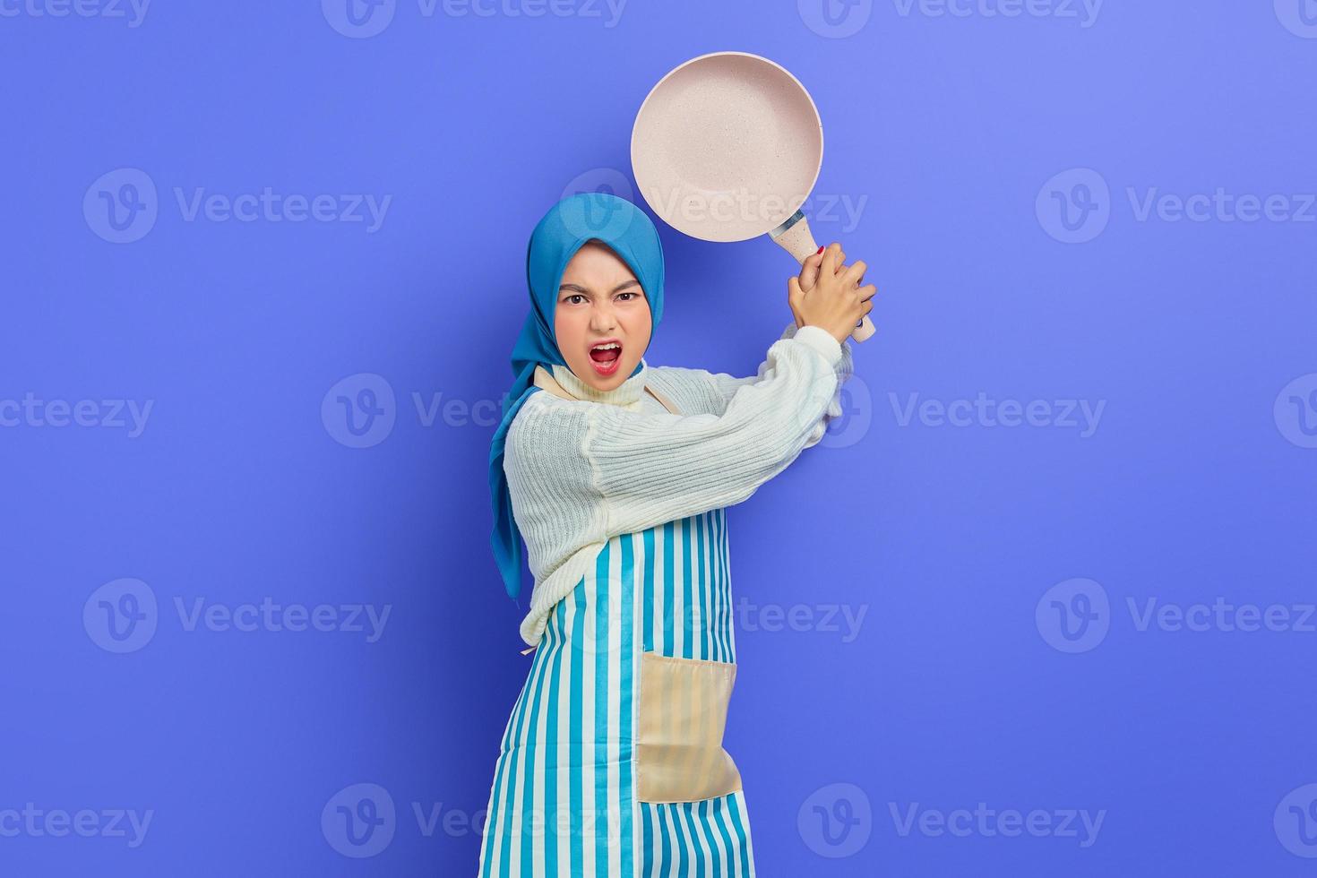 arrabbiata giovane donna asiatica in maglione bianco coperto di grembiule e hijab tenere la padella attaccare i nemici mentre fa i lavori domestici isolati su sfondo viola. concetto di pulizia foto