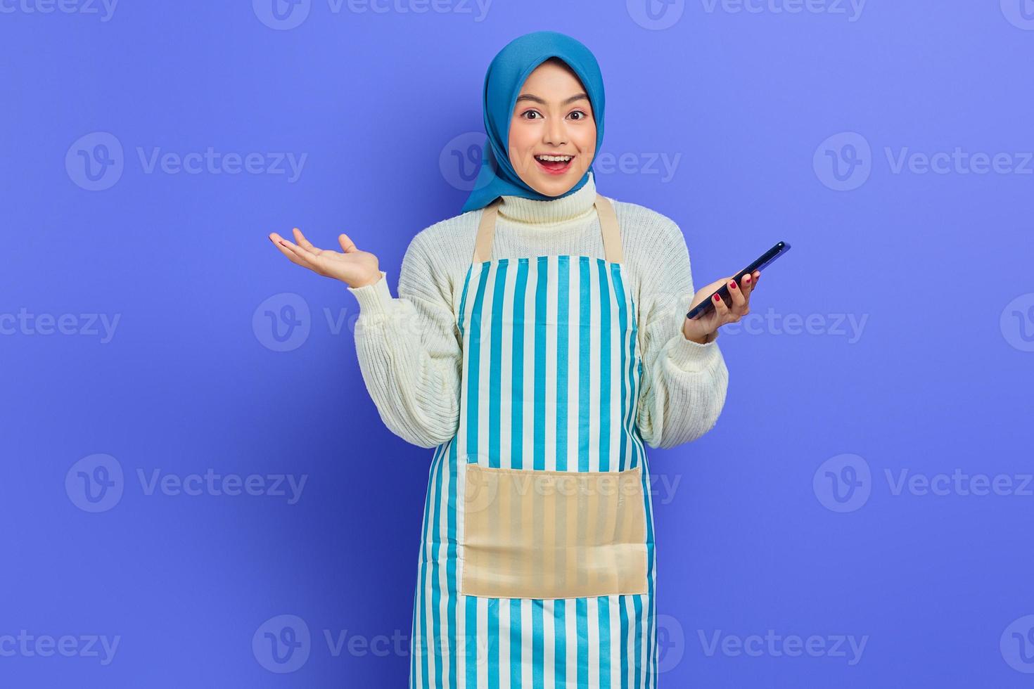 ritratto di eccitata giovane donna musulmana asiatica negli anni '20 che indossa hijab e grembiule utilizzando il telefono cellulare mentre guarda la fotocamera isolata su sfondo viola. concetto di stile di vita musulmano casalinga di persone foto