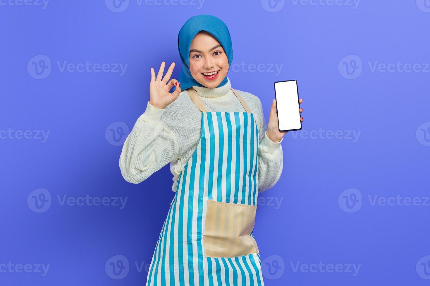 Ritratto di giovane donna musulmana asiatica sorridente negli anni '20 che indossa hijab e grembiule con in mano il telefono cellulare mentre mostra un gesto ok isolato su sfondo viola. concetto di stile di vita musulmano casalinga di persone foto